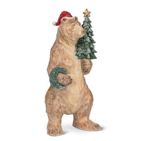 Wood look bear figurine wearing a Santa hat and holding a Christmas tree on a white background