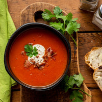 A bowl of tomato soup with a dollop of cream and crispy bacon on top, served on a wooden cutting board with parsley, next to a green napkin, a spoon, and slices of bread on a rustic wooden table.