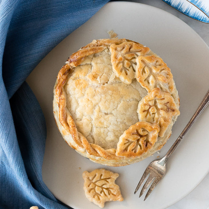 A baked toutiere pie with decorative leaf-shaped crust decorations sits on a white plate on a white marble surface, styled with a blue cloth, a fork, cookies, and blue-and-white ceramic mugs.
