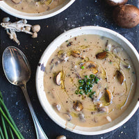Two bowls of creamy mushroom soup garnished with chives and olive oil, arranged on a dark wooden board with mushrooms, a spoon, and chives.