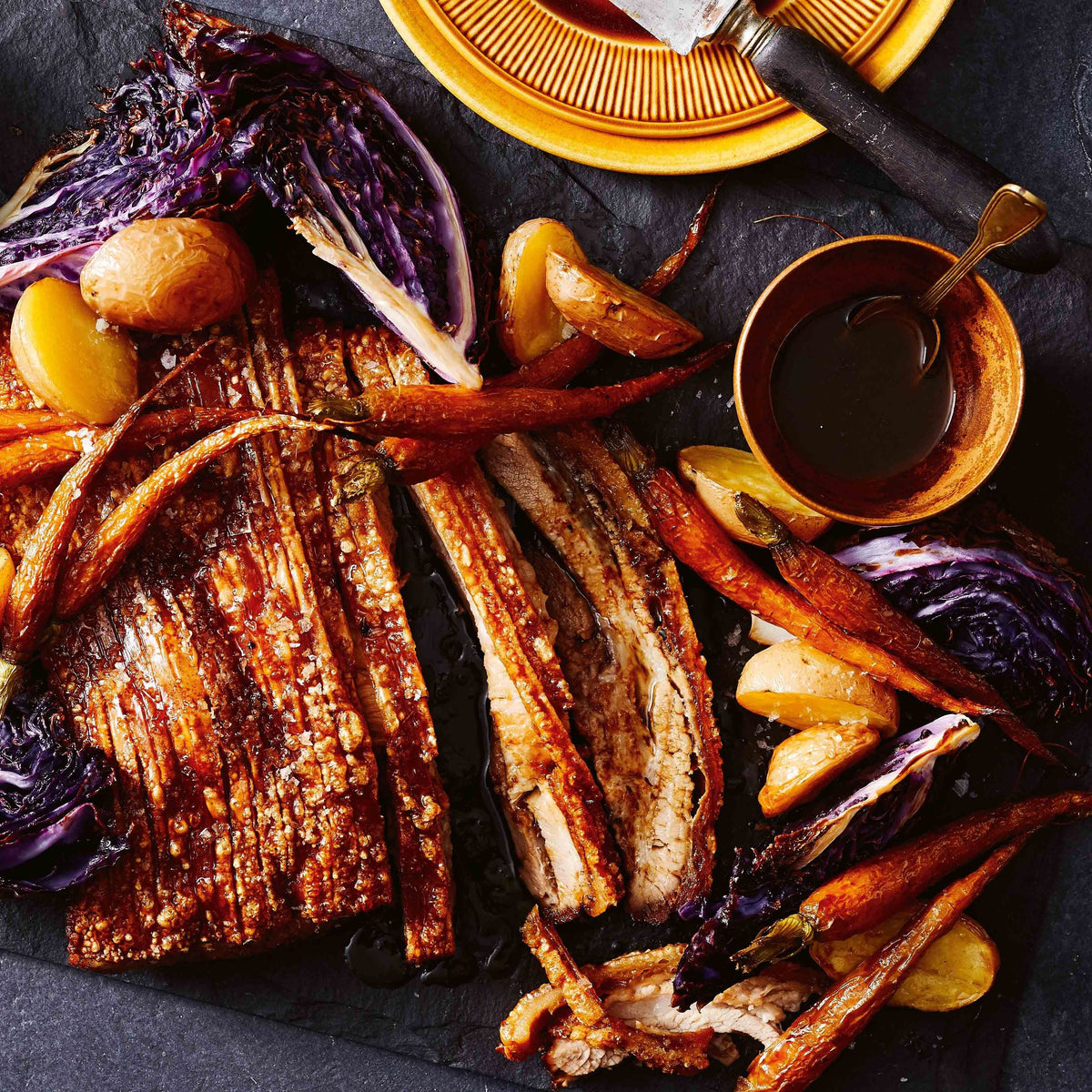 Overhead food photography of a cooked meat platter with sliced browned meat, roasted carrots, purple cabbage, potatoes, and dipping sauce on a dark slate surface.