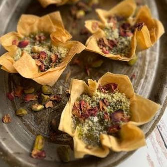 Six golden-brown puffy pastry cups filled with green pistachio and dried rose petals, arranged on a rustic oval metal platter.