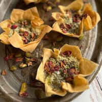 Six golden-brown puffy pastry cups filled with green pistachio and dried rose petals, arranged on a rustic oval metal platter.