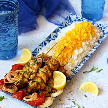 A rectangular blue-and-white ceramic platter holding skewered grilled chicken with tomatoes and onions, served over white rice topped with shredded yellow cheese, on a light surface with lemon wedges, herbs, and blue glass mason jars in the background.