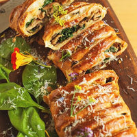 Crispy baked stuffed rolls or strudel slices on a wooden serving board, garnished with grated cheese, fresh spinach leaves, and edible flowers, with a knife visible at the top left on a light wood surface.