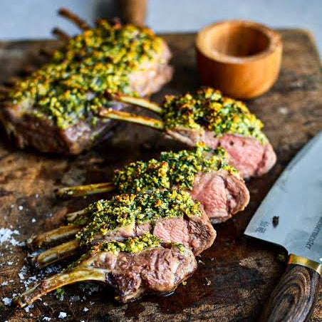 Three cooked lamb chops topped with a green herb sauce on a rustic wooden cutting board, with a chef's knife and a small wooden bowl nearby.