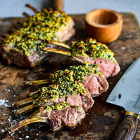 Three cooked lamb chops topped with a green herb sauce on a rustic wooden cutting board, with a chef's knife and a small wooden bowl nearby.