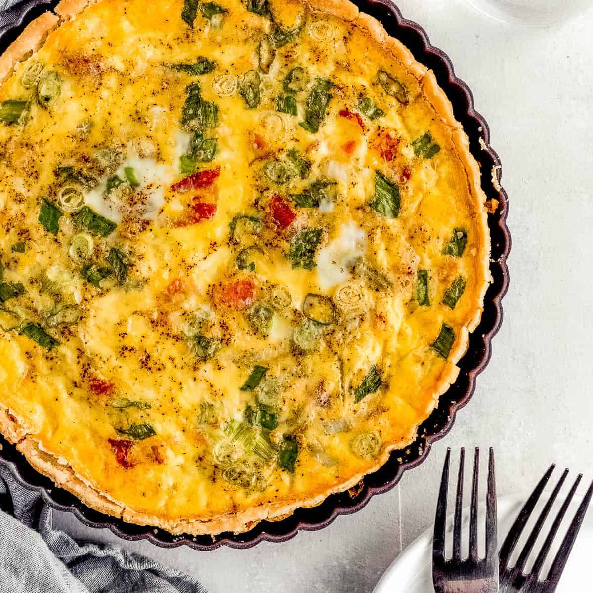 A baked quiche or savory pie with a golden yellow custard base, green onions, and small red tomato pieces, served in a dark fluted tart pan on a light gray surface with a glass of water, gray cloth, and forks nearby.