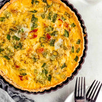 A baked quiche or savory pie with a golden yellow custard base, green onions, and small red tomato pieces, served in a dark fluted tart pan on a light gray surface with a glass of water, gray cloth, and forks nearby.