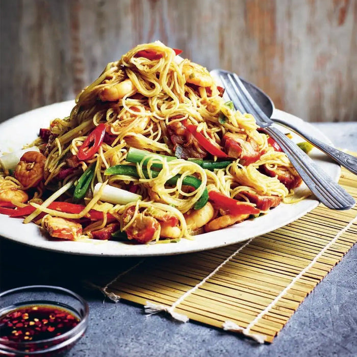 Plated dish of noodles with vegetables and meat on a textured surface, featuring 'The Curry Guy' branding.