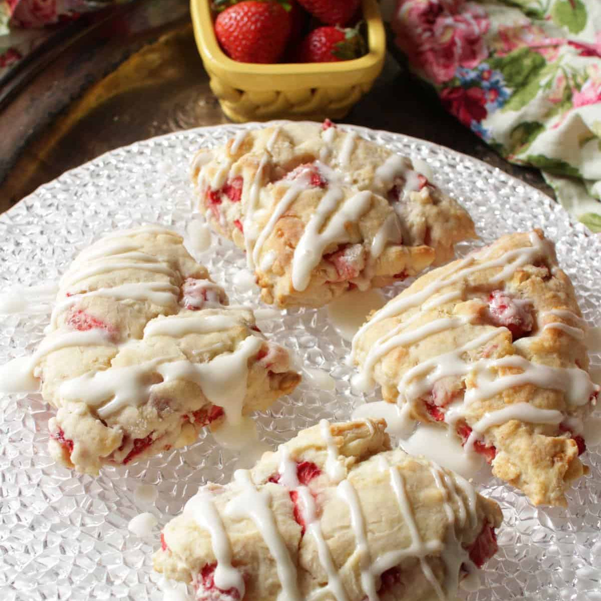 Four strawberry scones with white icing on a decorative plate, with strawberries in the background.