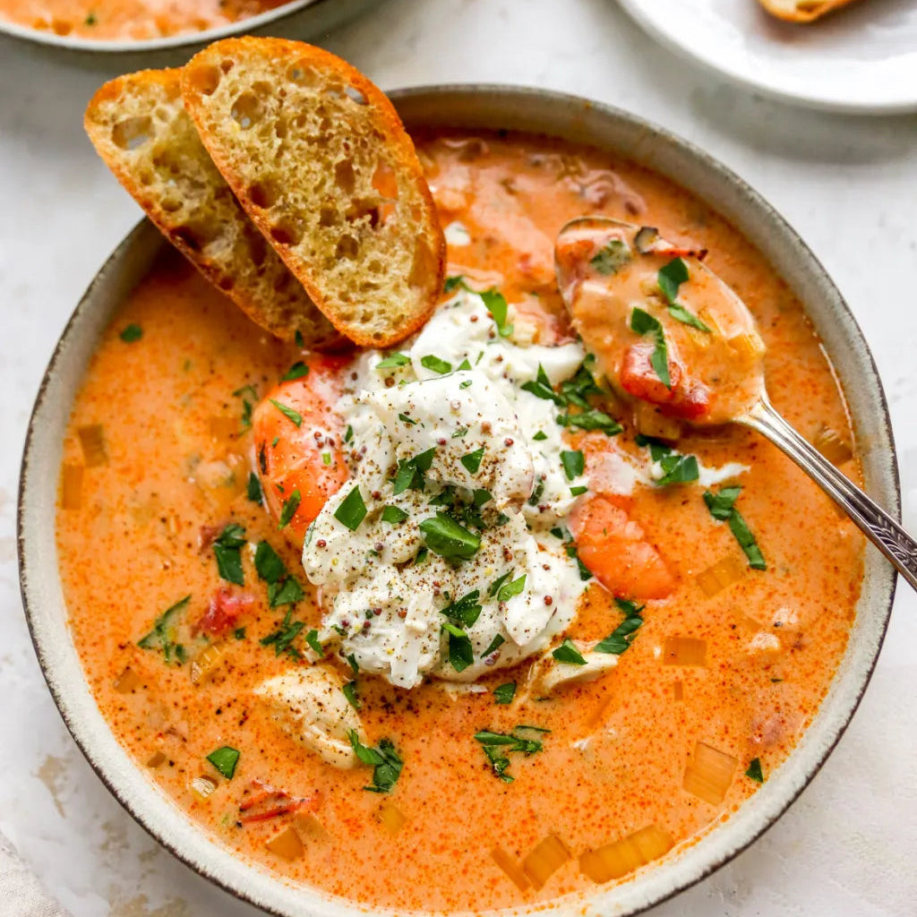 A bowl of creamy tomato, crap and shrimp soup topped with a dollop of white sour cream, chopped green herbs, and bread slices, photographed on a light tabletop with additional bread and a plate in the background.